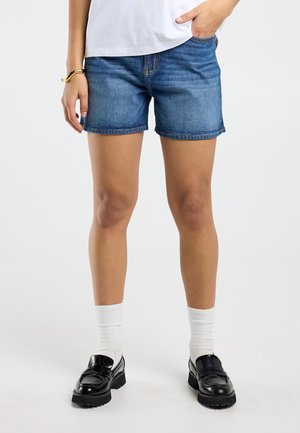 Persoon draagt middenblauwe denim shorts, witte sokken, zwarte loafers, wit shirt, met één hand in de zak en een gouden armband om de pols.