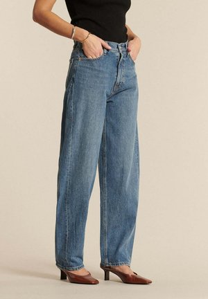 Kobieta ubrana w luźne niebieskie dżinsy z wysokim stanem, czarny top bez rękawów, brązowe buty na obcasie i złote bransoletki, stojąca z rękami w kieszeniach.