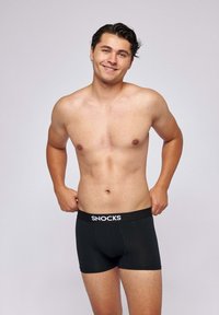 Young man smiling, shirtless, adjusting black boxer briefs with "SNOCKS" logo on waistband against plain background.