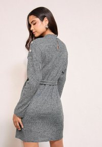 Abito grigio a maglia con maniche lunghe, vita aderente e dettaglio a chiave sulla schiena. Include una cintura in tessuto coordinato e bottoni decorativi sulle spalle.