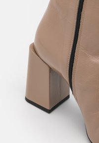 Bottine beige à talon bloc, avec une texture en cuir lisse et un détail zippé sur le côté. Base du talon carrée pour plus de stabilité.