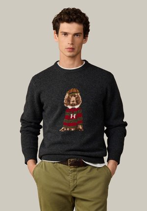 Joven con pelo rizado lleva un suéter oscuro con un perro con suéter a rayas rojas y verdes y gorra, combinado con pantalones verde oliva.