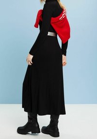 Schwarzes Langarmkleid mit einem fließenden Rock, kombiniert mit einem roten Pullover, der an den Schultern gebunden ist, und klobigen schwarzen Stiefeln. Silberne Gürteldetails.