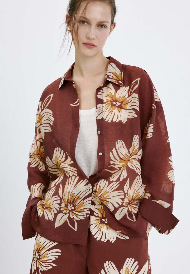 Femme portant un ensemble ample chemise et pantalon bordeaux avec de grands imprimés floraux beige et jaune sur un haut blanc, debout devant un fond uni.