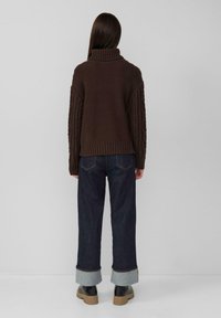 Brauner Strickpullover mit hohem Kragen und strukturierten Zopfmusterärmeln, kombiniert mit dunklen Jeans und schwarzen Stiefeletten.