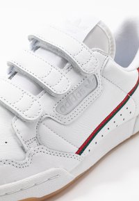 Vita lädersneakers med tre kardborreband, grön och röd accentrand och texturerad gummisula. Har en präglad logga på sidan.