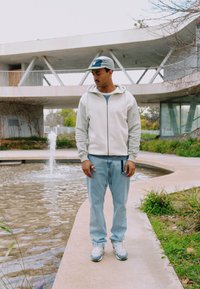 Homme portant une veste légère, une casquette et un jean, debout sur un chemin courbe près d’un étang avec une petite fontaine d’eau près d’un bâtiment moderne.