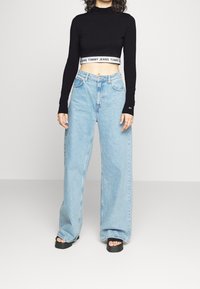 Svart långärmad croppad topp med vit logoband och ljusblå vida jeans. Svarta platåsandaler kompletterar outfiten.