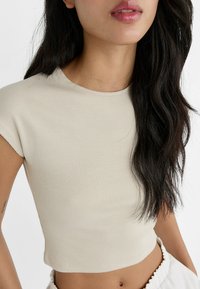 Beżowy prążkowany crop top z krótkimi rękawami i okrągłym dekoltem, o miękkiej fakturze i dopasowanym kroju, w połączeniu z jasnymi spodniami.