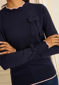 Námořnický modrý pletený svetr se zaobleným růžovým dekoltem a manžetami. Má mašličku na hrudi; měkká textura, přiléhavý střih.