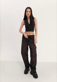 Kvinne med langt mørkt hår, iført en sorte ermeløs zip-up crop top, baggy sorte jeans og sorte plattformsko, stående mot en ensfarvet baggrund.