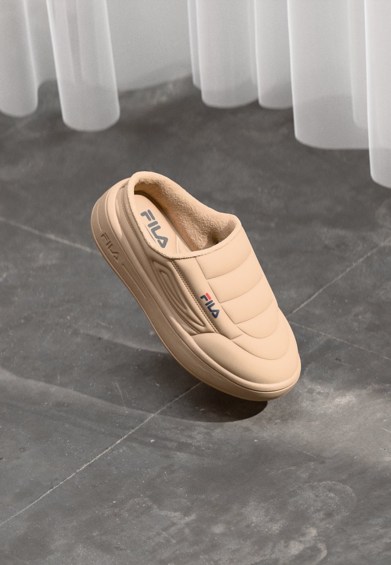 Beige FILA slip-on sko med polstret overdel og blødt foer, placeret på en grå flisegulv under gennemsigtige hvide gardiner.