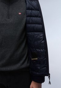 Uomo che indossa un maglione grigio scuro con una piccola toppa rossa-bianca-blu e una giacca a vento nera aperta che mostra la tasca interna con il marchio e le linguette della cerniera.