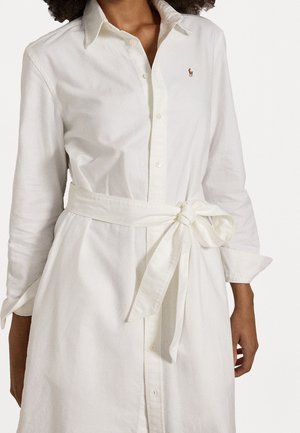 Femme portant une robe blanche à manches longues boutonnée avec une ceinture en tissu nouée et un petit logo brodé sur la poitrine.