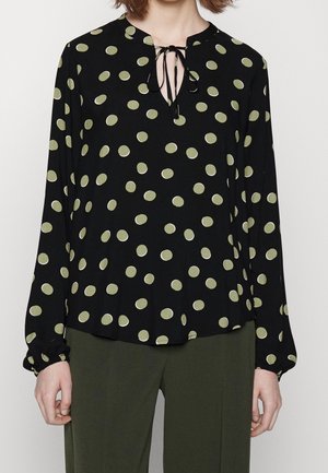 Femme portant un chemisier noir à manches longues avec des pois verts et un décolleté en goutte noué avec un cordon noir, assorti à un pantalon vert olive.