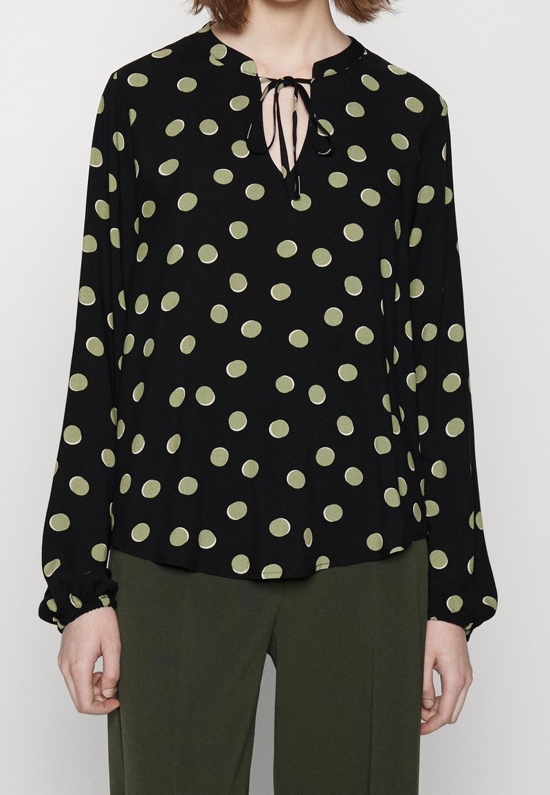 Femme portant un chemisier noir à manches longues avec des pois verts et un décolleté en goutte noué avec un cordon noir, assorti à un pantalon vert olive.