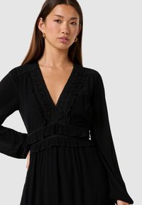 Vestito nero con maniche lunghe, scollatura a V profonda, dettagli arricciati e decorazioni in pizzo lungo il collo e la vita, realizzato in tessuto testurizzato.