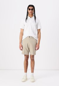 White polo shirt with logo, beige shorts, and cream sneakers. Model wears dark sunglasses, stands against a plain white background.