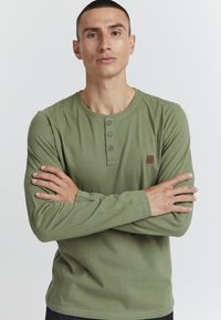 Olivovo zelená dlhá rukávová henley košeľa s trojbuttonovou légou, malou logom na hrudi a viditeľnými švovými detailmi.
