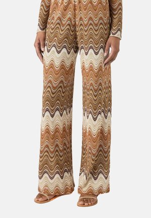 Pantaloni a gamba larga con motivo ondulato a zigzag nei toni del marrone, beige e arancione, indossati con sandali aperti color cuoio su sfondo chiaro.