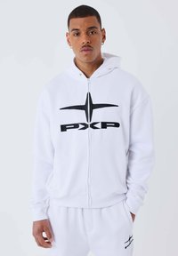 Sudadera blanca con cremallera en tejido suave, con un gran logo negro "PXP" y una línea de acento en el frente, que cuenta con una capucha forrada y puños de canalé.