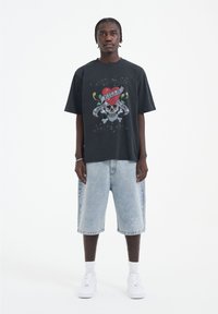 Černé grafické tričko s designem lebky a srdce, spárované se světle modrými denimovými šortkami. Bílé tenisky dotvářejí vzhled.