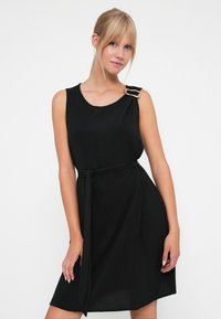 Robe noire sans manches avec une silhouette ajustée, un tissu texturé et un détail de taille nouée. Présente un accent métallique sur une épaule.