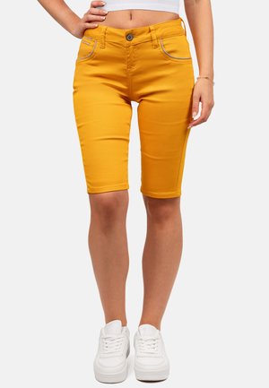 Vrouw draagt mosterdgele knielange shorts, witte sneakers en een wit crop top, staand met hand op heup.