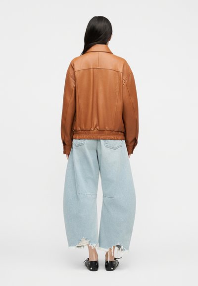 Γυναίκα που φαίνεται από πίσω, φοράει καφέ δερμάτινο μπουφάν, ανοιχτό μπλε φαρδιά σκισμένα τζιν και μαύρα παπούτσια με τρουκς, σε λευκό φόντο.