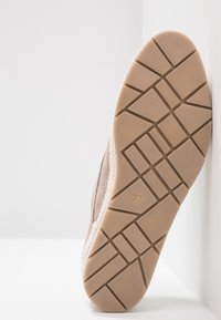 Semelle d'une basket beige montrant un motif de traction géométrique et la pointure 37, appuyée contre un coin blanc.