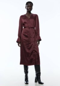 Bordeaux Satin-Wickelkleid mit langen Ärmeln und einem Bindegürtel, mit vorderen Knöpfen und geradem Schnitt, ergänzt durch knielange schwarze Stiefel.
