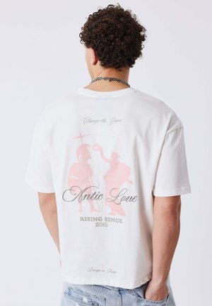 Homme aux cheveux bouclés portant un T-shirt blanc avec une illustration rose d'un guerrier grec classique et d'une femme, accompagnée du texte « Amour Antique, en ascension depuis 2015 ».