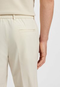 Man i beige perforerade byxor med elastisk midja och en horisontell bakficka, stående med handen avslappnad vid sidan.