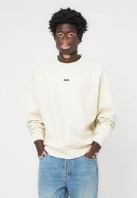 Sudadera color crema con cuello redondo, mangas largas, puños acanalados y un pequeño logo "BOSS" en negro en el pecho. Llevada con vaqueros azules.