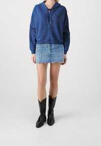 Sudadera azul con cremallera y acabado texturizado, combinada con una minifalda de mezclilla con bordes deshilachados y costuras en contraste, y botas vaqueras negras.