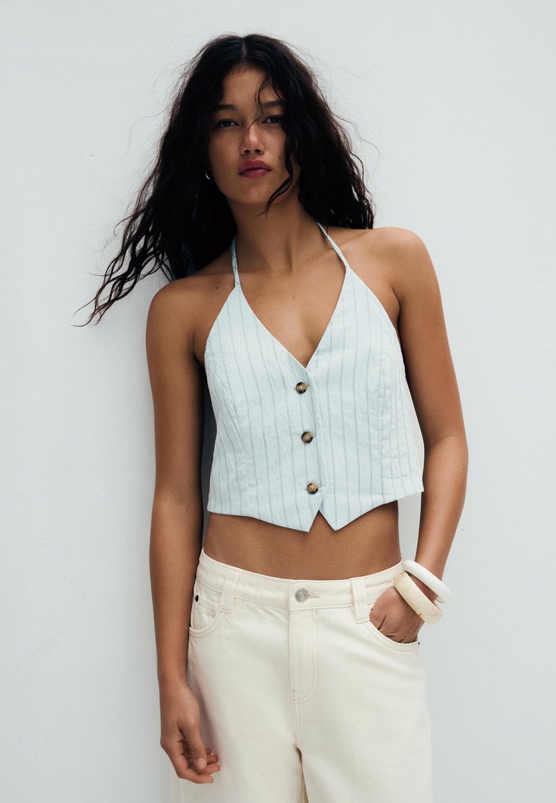 Jeune femme aux longs cheveux bouclés portant un haut bleu clair à col halter avec des boutons et un pantalon crème, debout devant un fond clair uni.