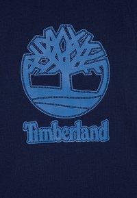 Camiseta azul marino con un gran logo del árbol de Timberland en azul y texto, ambos con un diseño de contorno bordado. Material de algodón con textura suave.