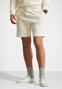 Ljust gula bomullsshorts med elastisk midja, dragsko, sidofickor och en avslappnad passform, kombinerade med grå strumpor och vita gympaskor.
