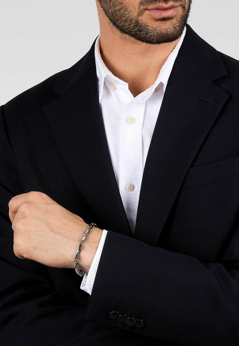 Silverkedja armband med ihopkopplade länkar, buren på ett handled. Svart kavaj och vit skjorta med knappdetaljer är synliga.