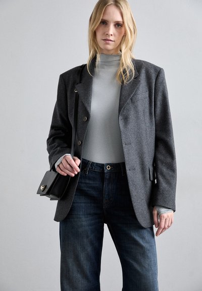 Plavolasa ženska nosi temnosivo oversize sakou, svetlosivo pulover s turtleneckom, modre kavbojke in drži črno torbico čez ramo.