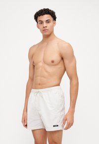 Costumi da bagno color beige chiaro con elastico in vita e cordoncino. Presentano tasche laterali e un piccolo etichetta nera sul bordo. Tessuto liscio.