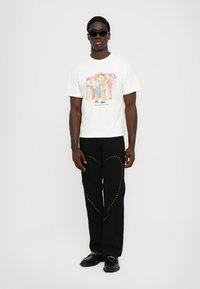 White short-sleeve t-shirt with colorful printed design, paired with black pants featuring gold studded heart pattern and black shoes.