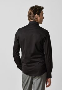 Zwarte lange mouwen shirt met een gladde textuur, klassieke kraag en knoopmanchetten, gecombineerd met grijze broek, gezien van achteren.