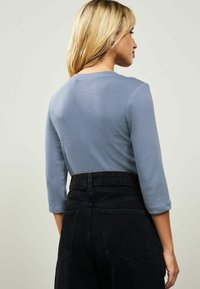 Femeie cu părul blond îmbrăcată într-o bluză de lungă cu mânecă lungă de culoare albastră deschisă și jeans negri din denim cu talie înaltă, stând de profil pe un fundal simplu.