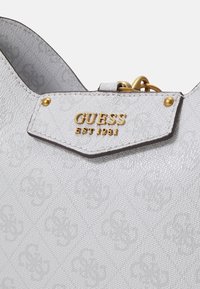 Sac à main blanc Guess avec motif embossé, plaque logo dorée indiquant "GUESS EST 1981" et accessoires métalliques dorés.