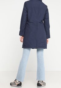 Mörkblå lång kappa med en slät yta, två sidofickor och en ryggslits, kombinerad med ljusblå utsvängda jeans och grå skor.