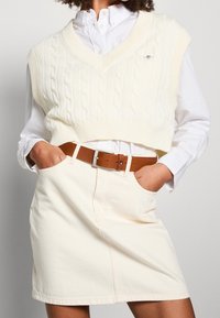 Personne portant un pull sans manches blanc en maille torsadée par-dessus une chemise blanche boutonnée, une jupe beige et une ceinture marron, les mains dans les poches.