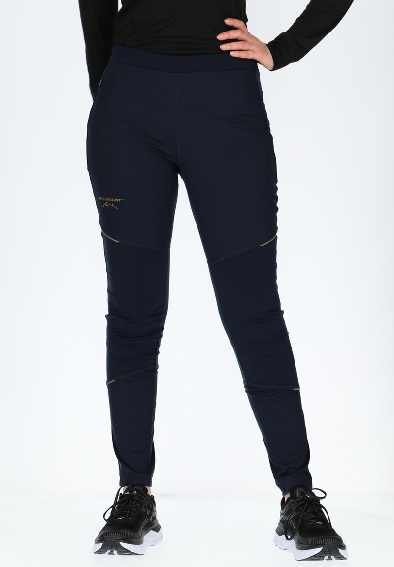 Pantaloni sportivi elasticizzati blu navy con vita alta, pannelli laterali texture e cuciture di accentuazione dorate. Indossati con scarpe da ginnastica nere.