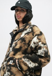 Casaco de pelúcia castanho e creme com um fecho de pressão preto e um emblema; apresenta um corte descontraído e um tecido suave e texturizado. Modelo com boné preto.