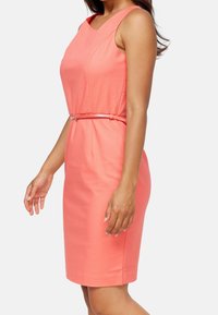 Korálové bezrukávové šaty s přiléhavým střihem, páskem v užší části pasu a jemnou tkaninou. Jednoduchý výstřih a délka po kolena.